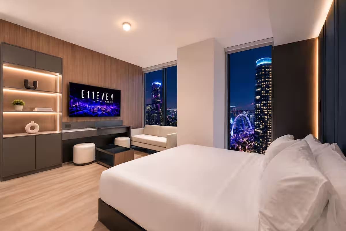 Bed beside city-view windows at dusk.