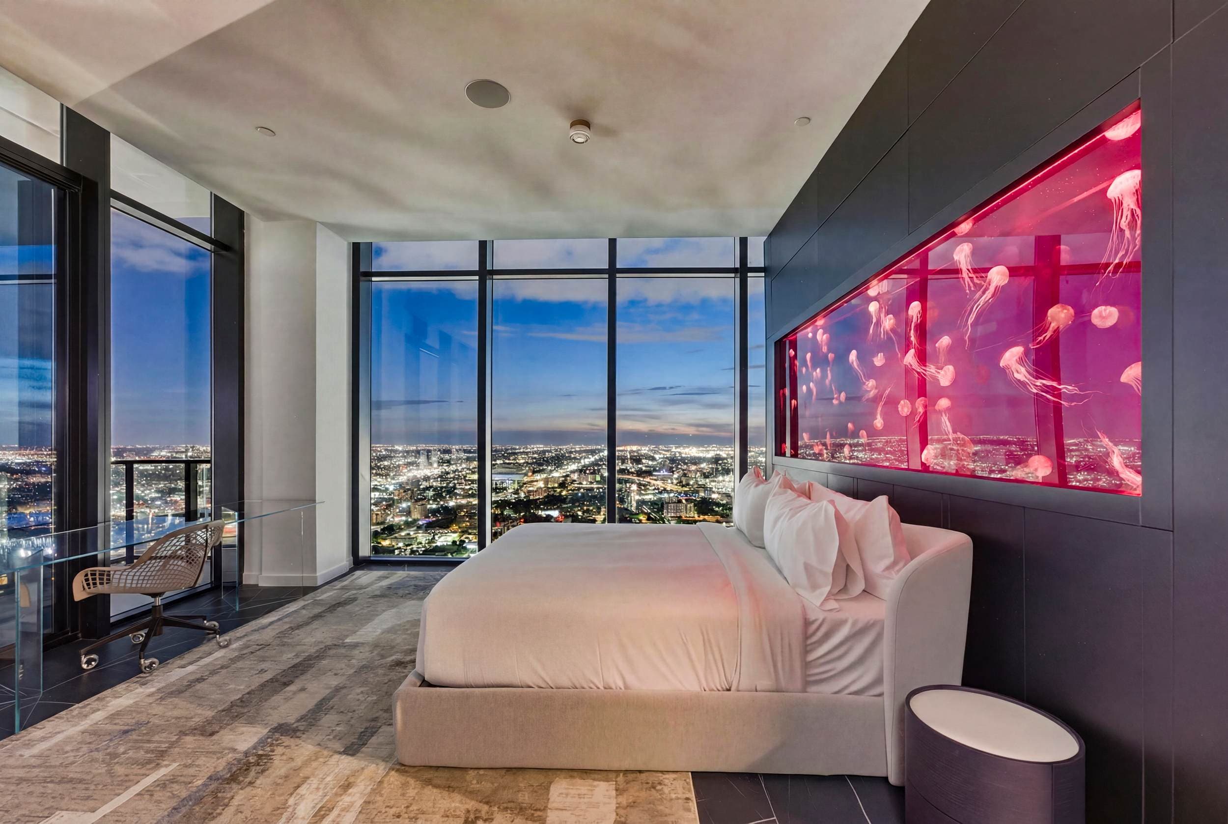 Bed beside illuminated pink artwork and city views.
