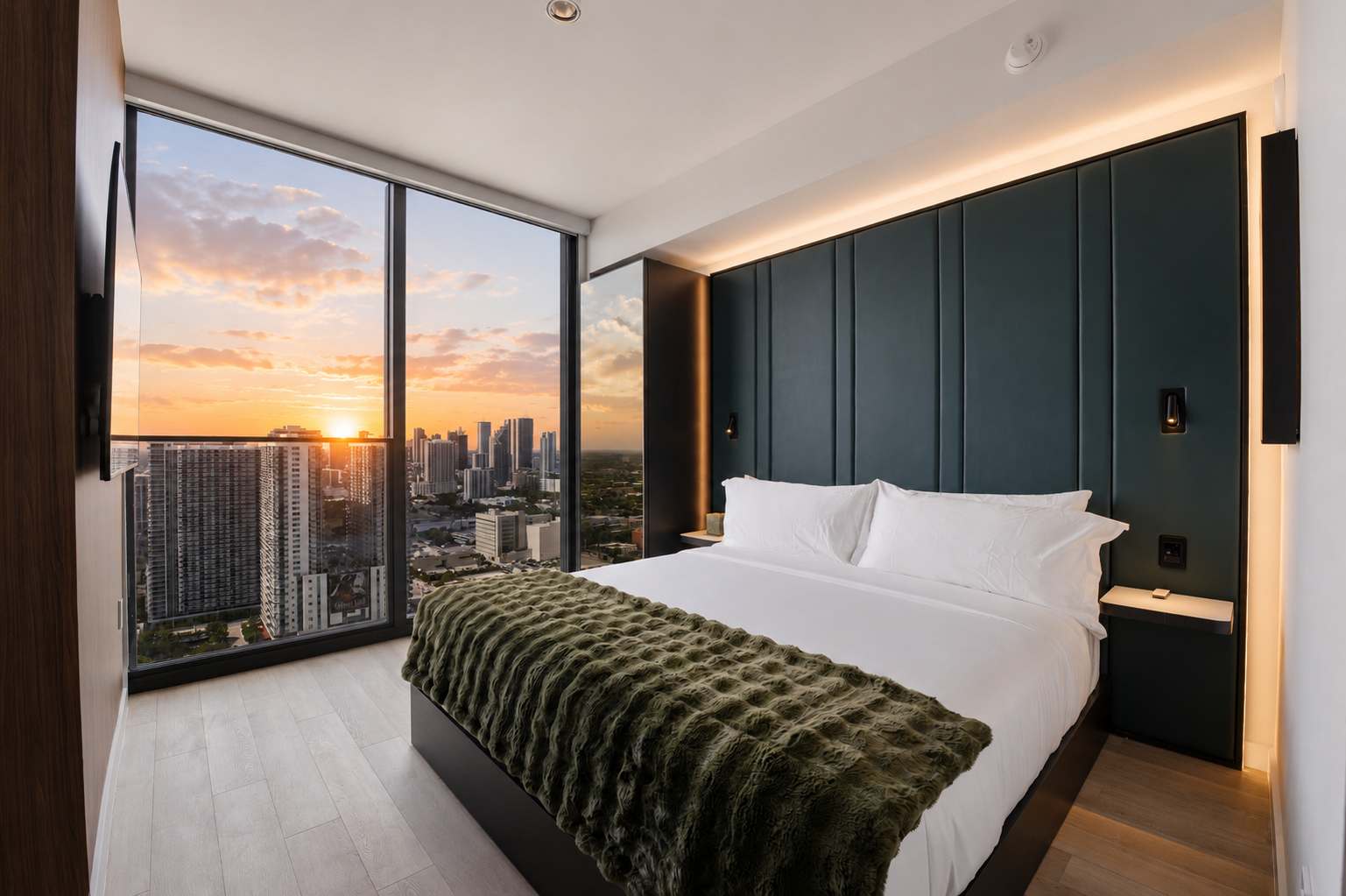 Bed beside city-view windows at dusk.