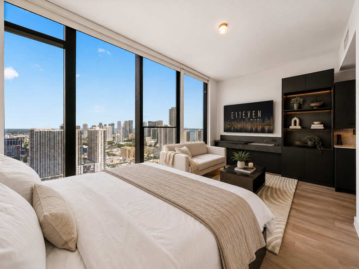 Bed with a beige throw beside city-view windows.