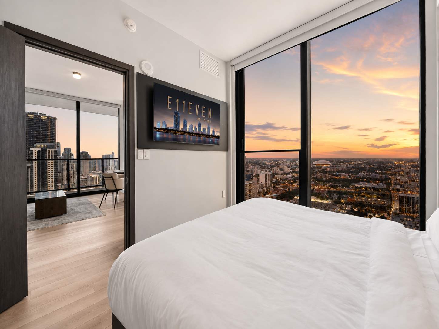 Bed beside city-view windows at dusk.
